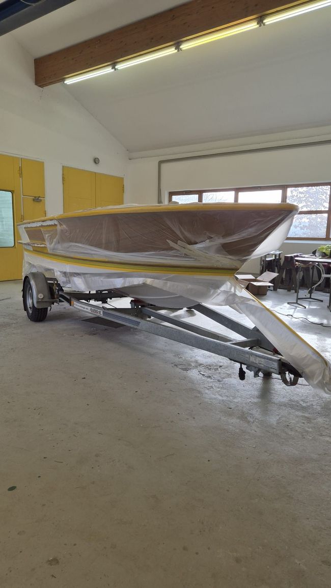 Ein eingewickeltes Boot auf einem Anhänger in einer Garage, mit gelben Schränken und Neonröhren an der Decke.