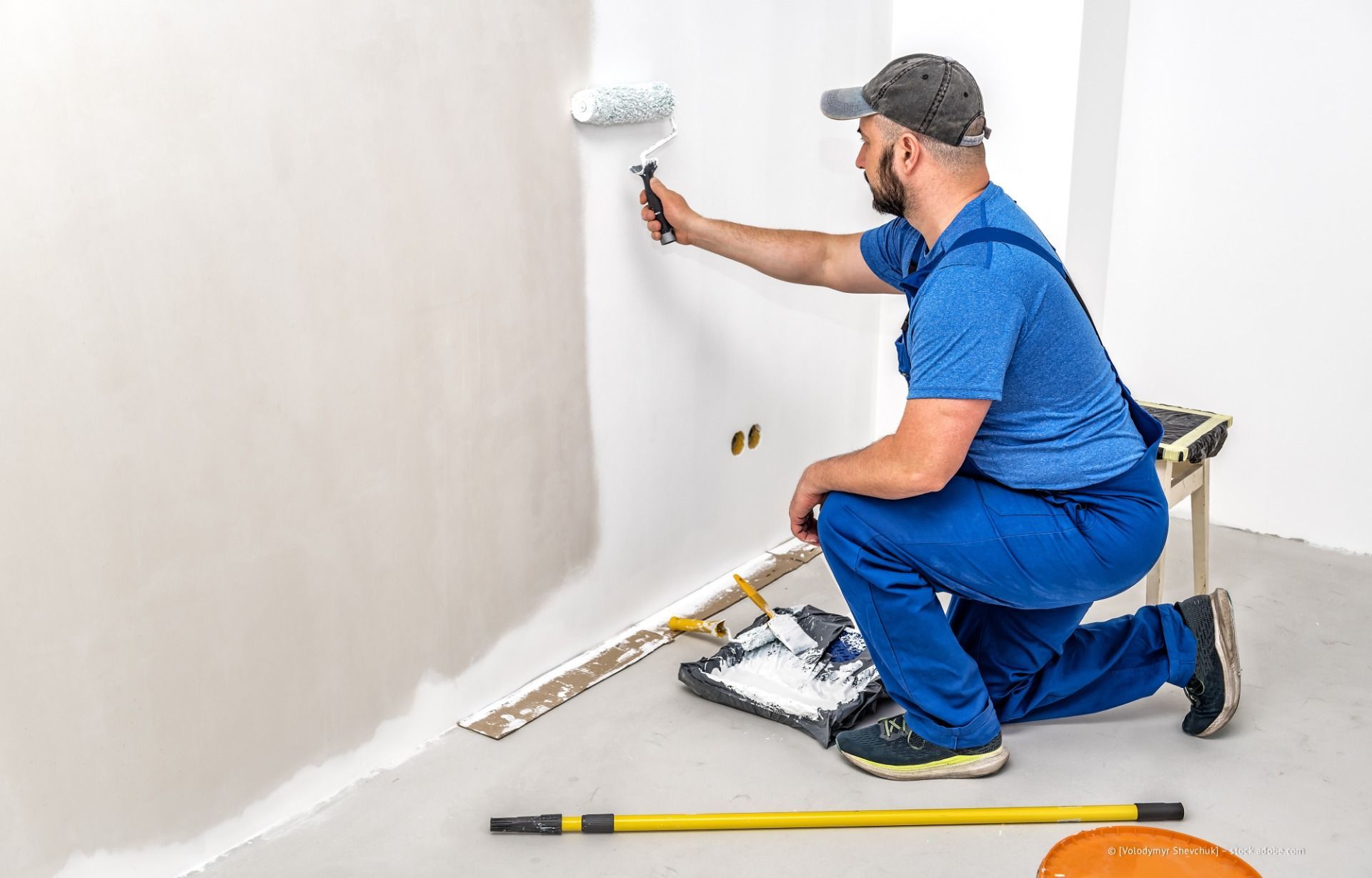 Maler im blauen Hemd kniet auf einer Abdeckplane und trägt weiße Farbe auf eine Wand auf.