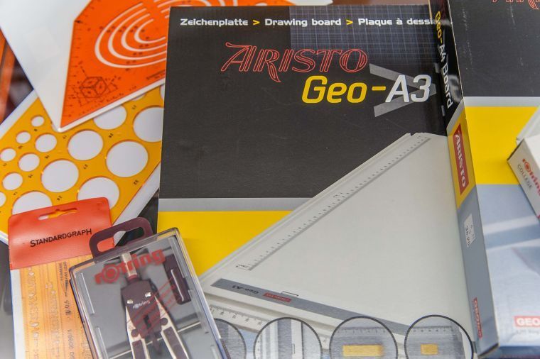 Verschiedene Zeichenmaterialien und Verpackungsmaterialien, darunter eine ARISTO Geo-A3-Box und farbige Ordner.