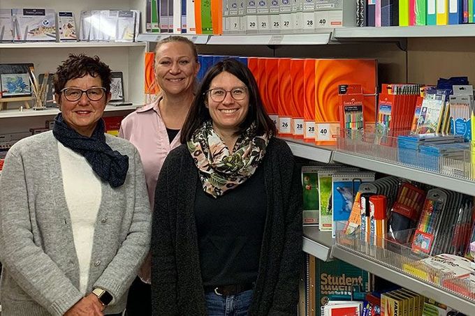 Drei lächelnde Personen stehen in einem Buchladengang, hinter ihnen Regale mit Büchern und Schreibwaren.
