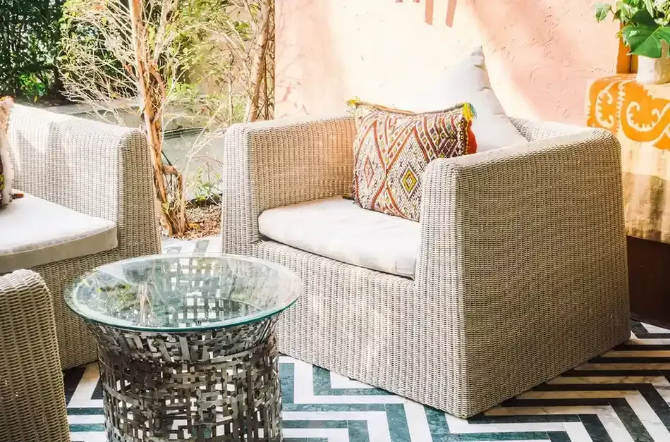 Sillas de mimbre para patio con cojines blancos y una mesa auxiliar con tapa de cristal sobre una alfombra.