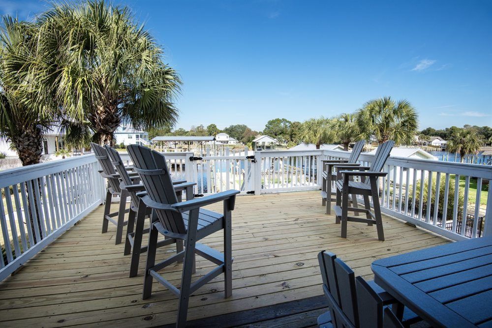 Wooden deck with bar stools, palm trees, and water view under a blue sky.