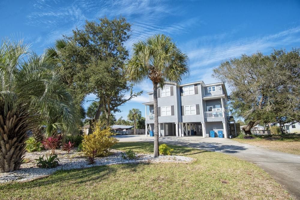 Multi-story gray house with balconies, palm trees, and driveway on a sunny day.