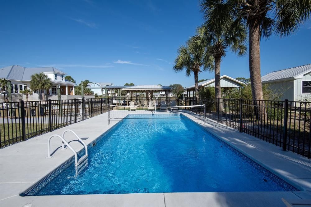 A rectangular pool with blue water, surrounded by concrete, a black fence, and palm trees under a blue sky.