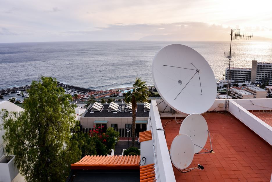 Tres antenas parabólicas en una azotea miran al océano y a una ciudad costera bajo un cielo nublado.