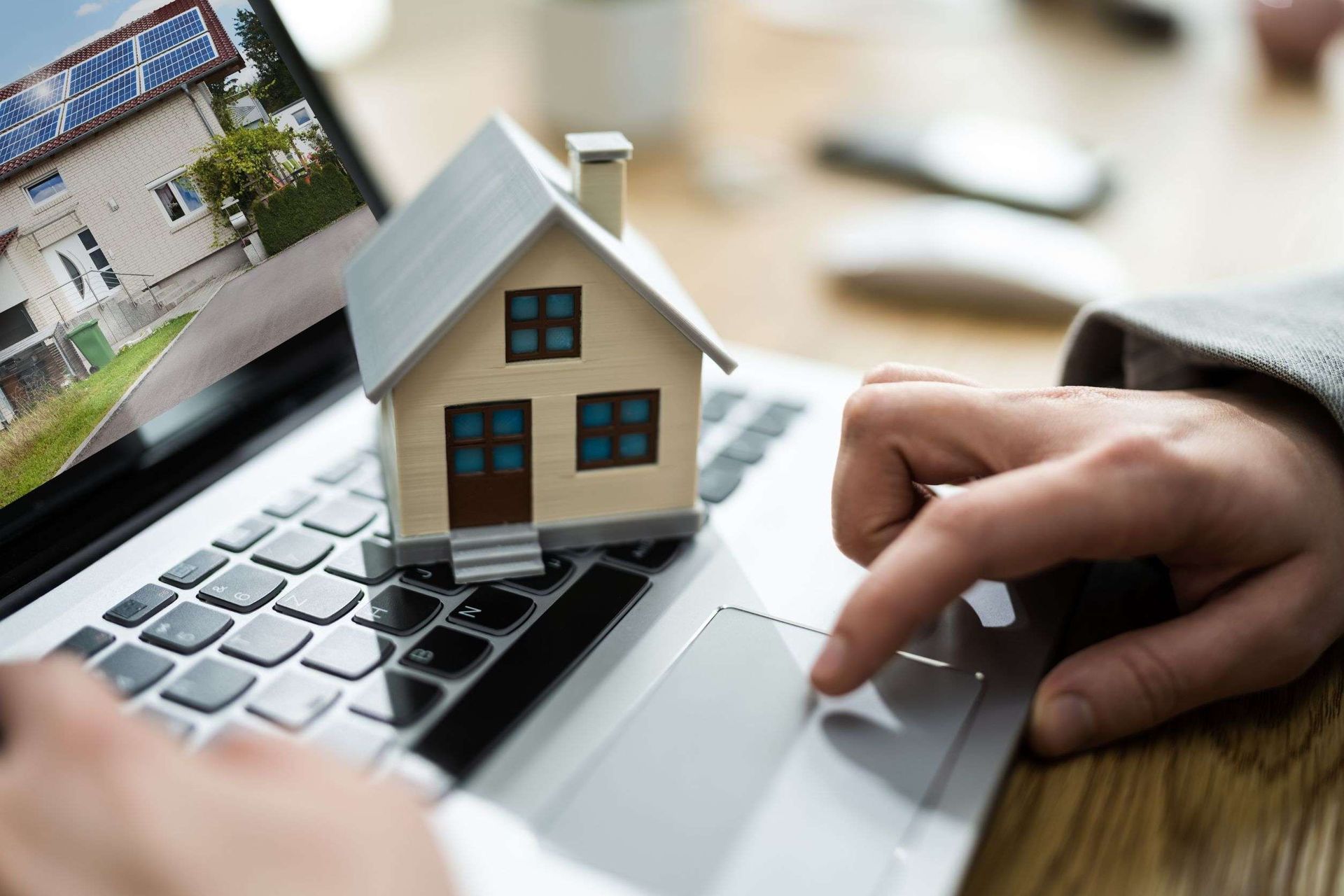 Persona que usa una computadora portátil con una casa en miniatura en el teclado y una imagen de una casa en la pantalla.