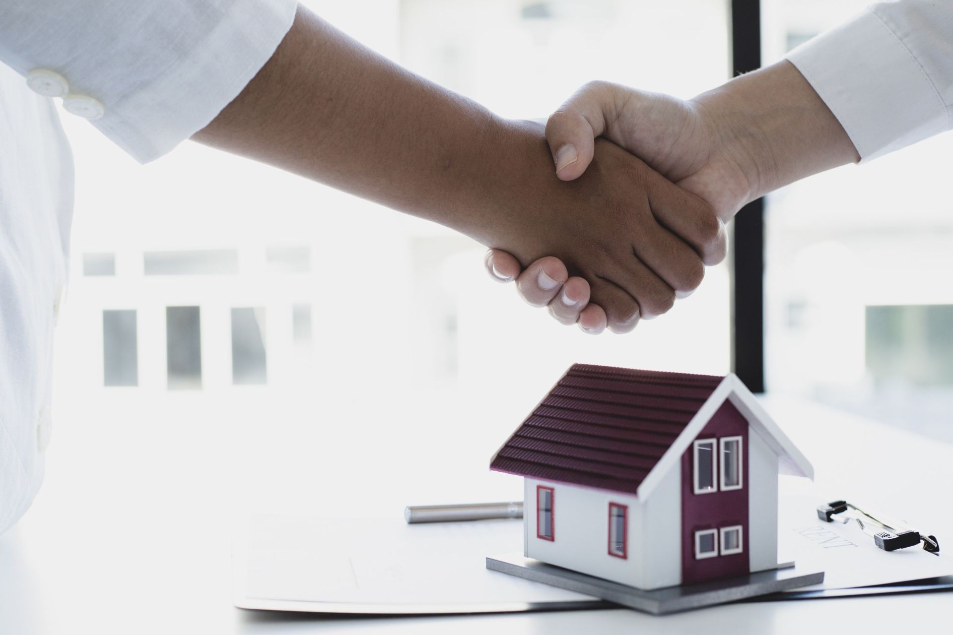 Dos personas se dan la mano sobre una casa en miniatura sobre una mesa.