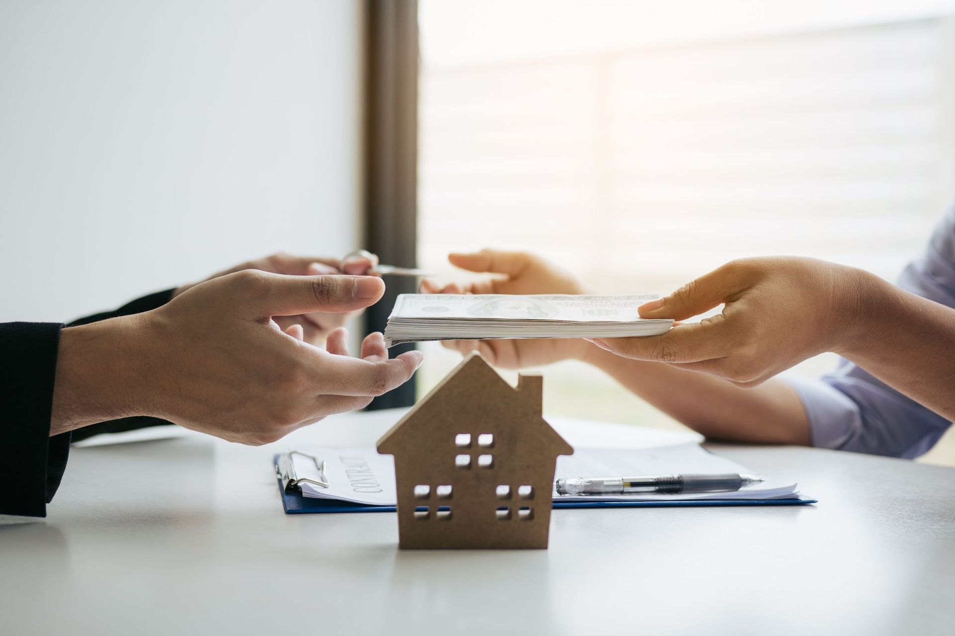 Una persona entrega dinero en efectivo a otra persona sobre una casa en miniatura sobre una mesa.