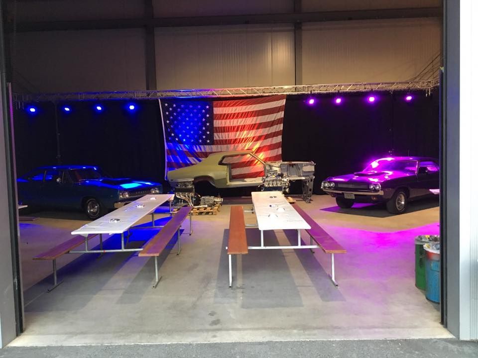 Im Hintergrund eine amerikanische Flagge, Oldtimer, Picknicktische in einem großen Innenraum mit blauer und rosa Beleuchtung.