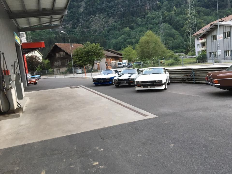 Drei Oldtimer parken in der Nähe einer Zapfsäule, im Hintergrund sind Berge zu sehen.