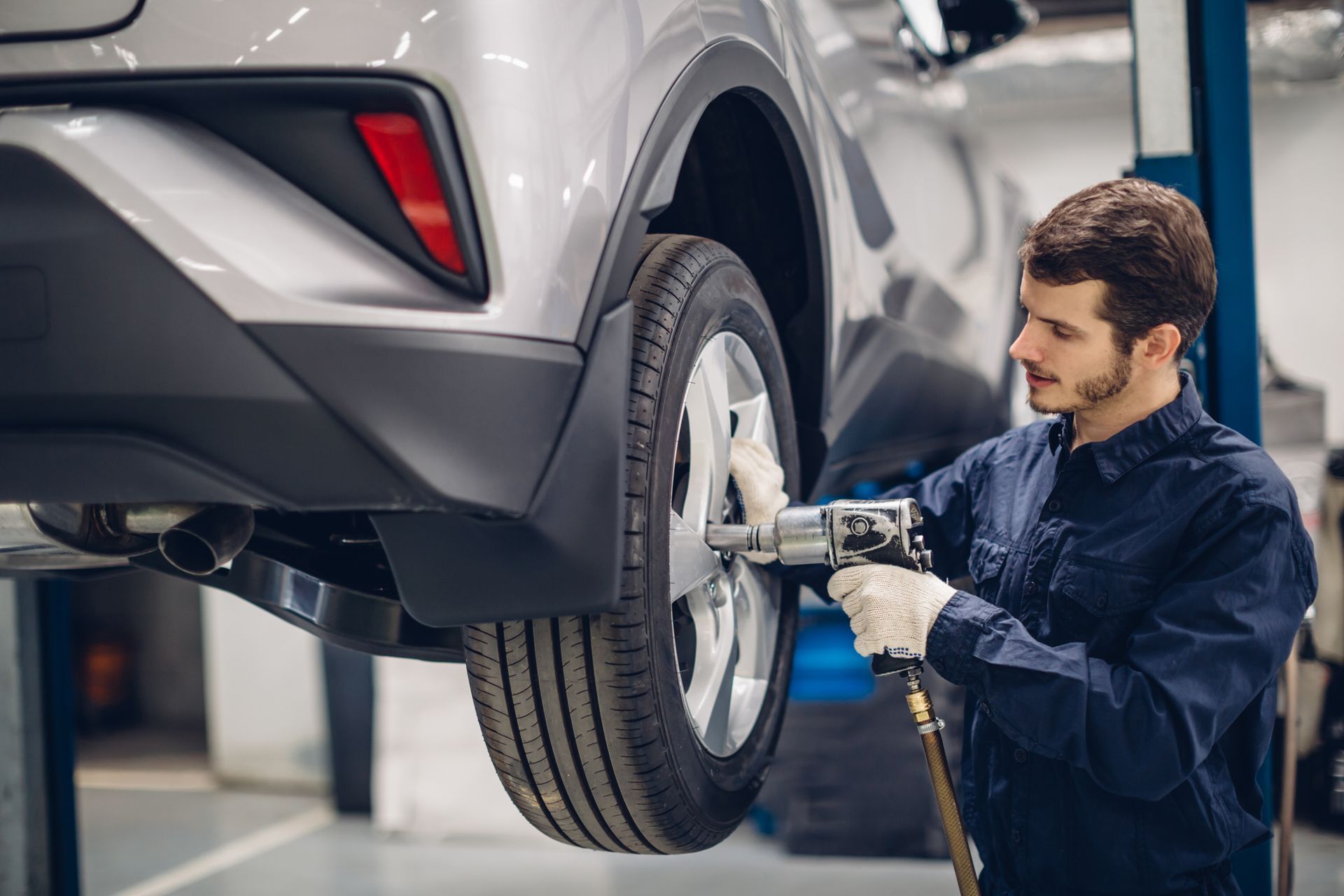Mechaniker in blauer Uniform, der in einer Garage einen Druckluftschrauber an einem Autoreifen benutzt.