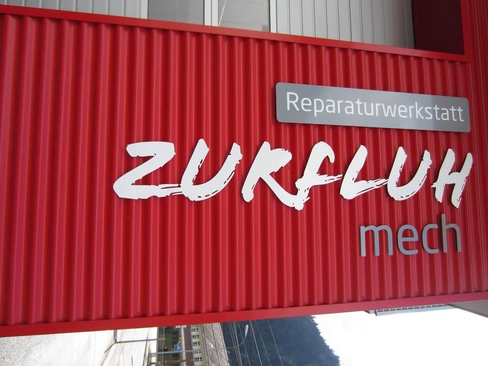 Rotes Wellblechschild mit weißem Text: ZURFLUH, darunter „mech“. Silbernes Schild: Reparaturwerkstatt.