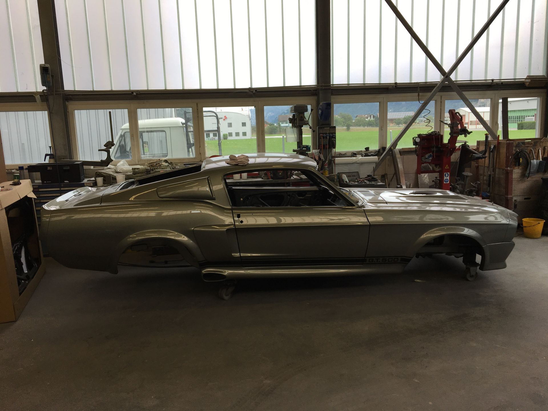 Ein silberner klassischer Ford Mustang wird in einer Garage restauriert; im Hintergrund sind Fenster zu sehen.