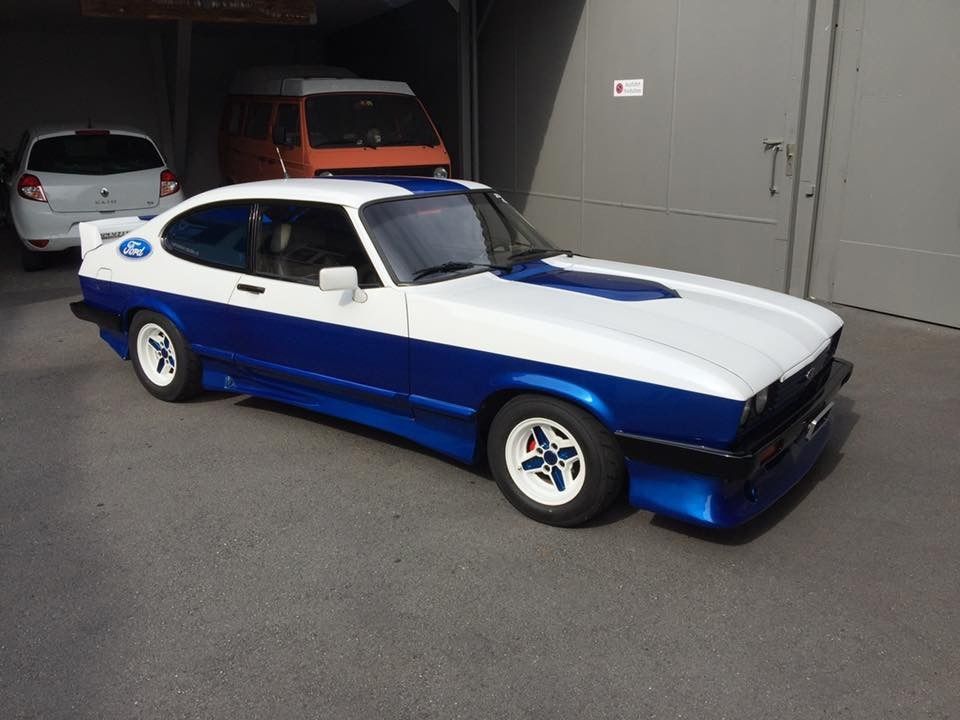 Blau-weißer Oldtimer, in der Nähe einer Garage geparkt; orangefarbener Lieferwagen und weißes Auto im Hintergrund.