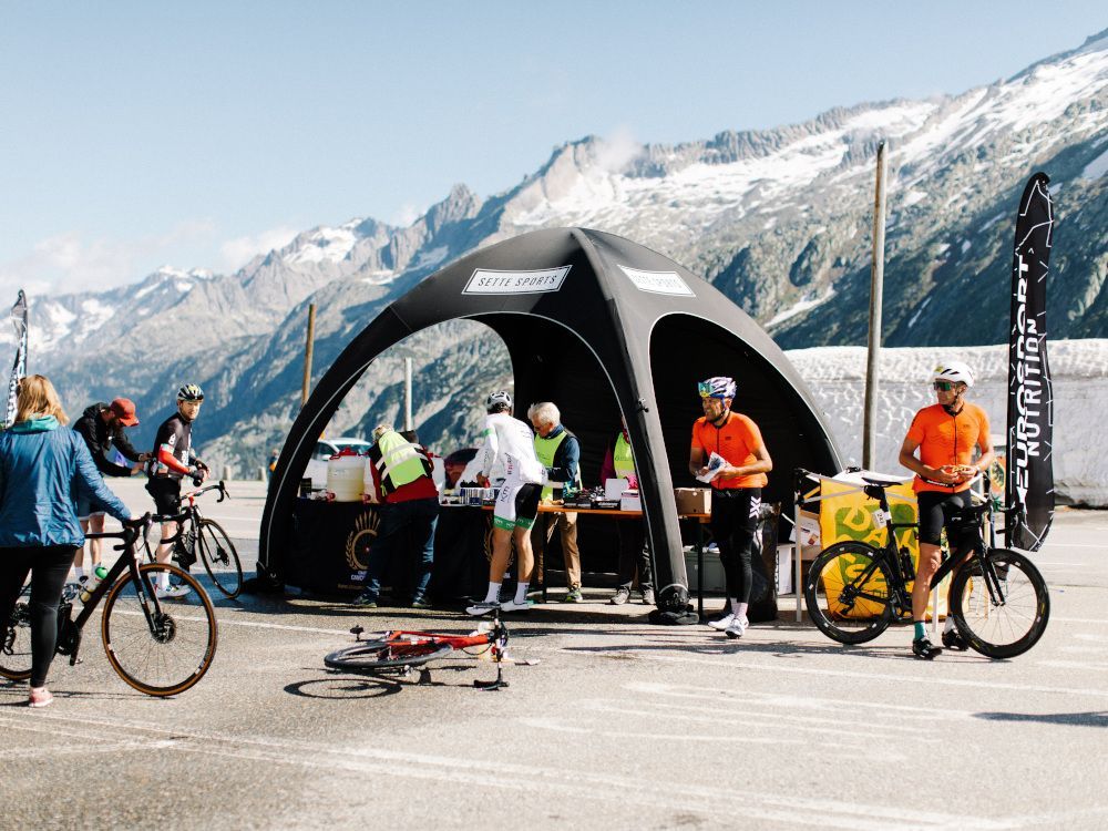 Cyclists and a rescue tent at a mountain road event with snowy peaks in the background