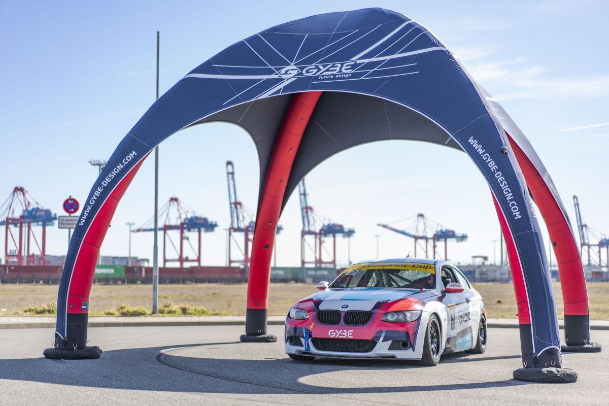 Race car under a blue arch at a waterfront track, with cranes and sea in the background