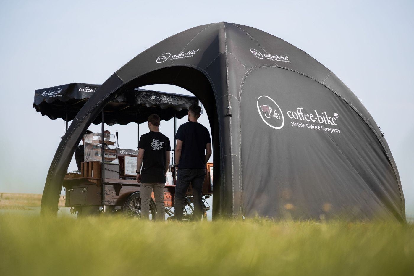 Two people standing by a dark camping tent in a grassy field.