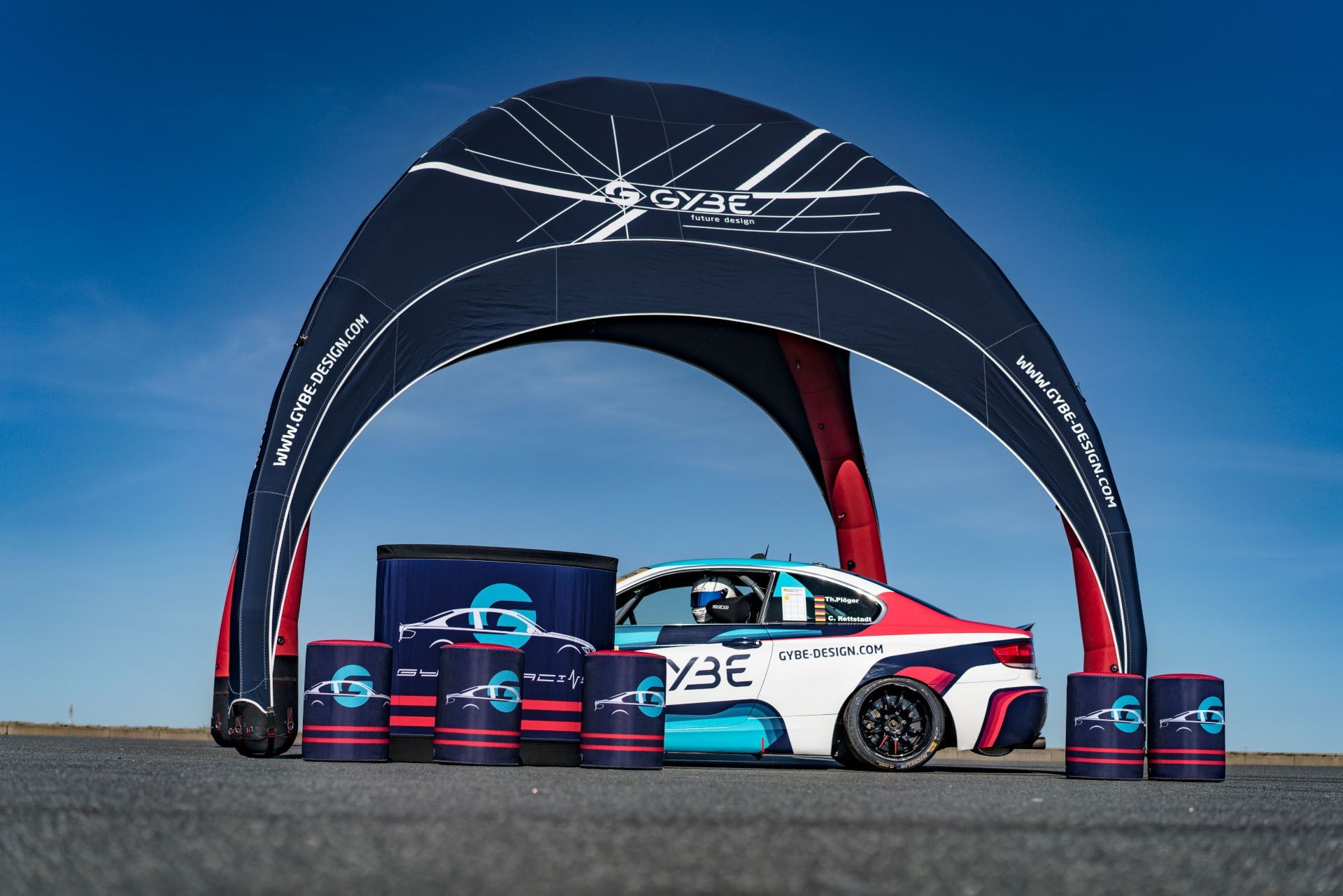 Race car under a black canopy with stacked sponsor barrels on a track under a blue sky