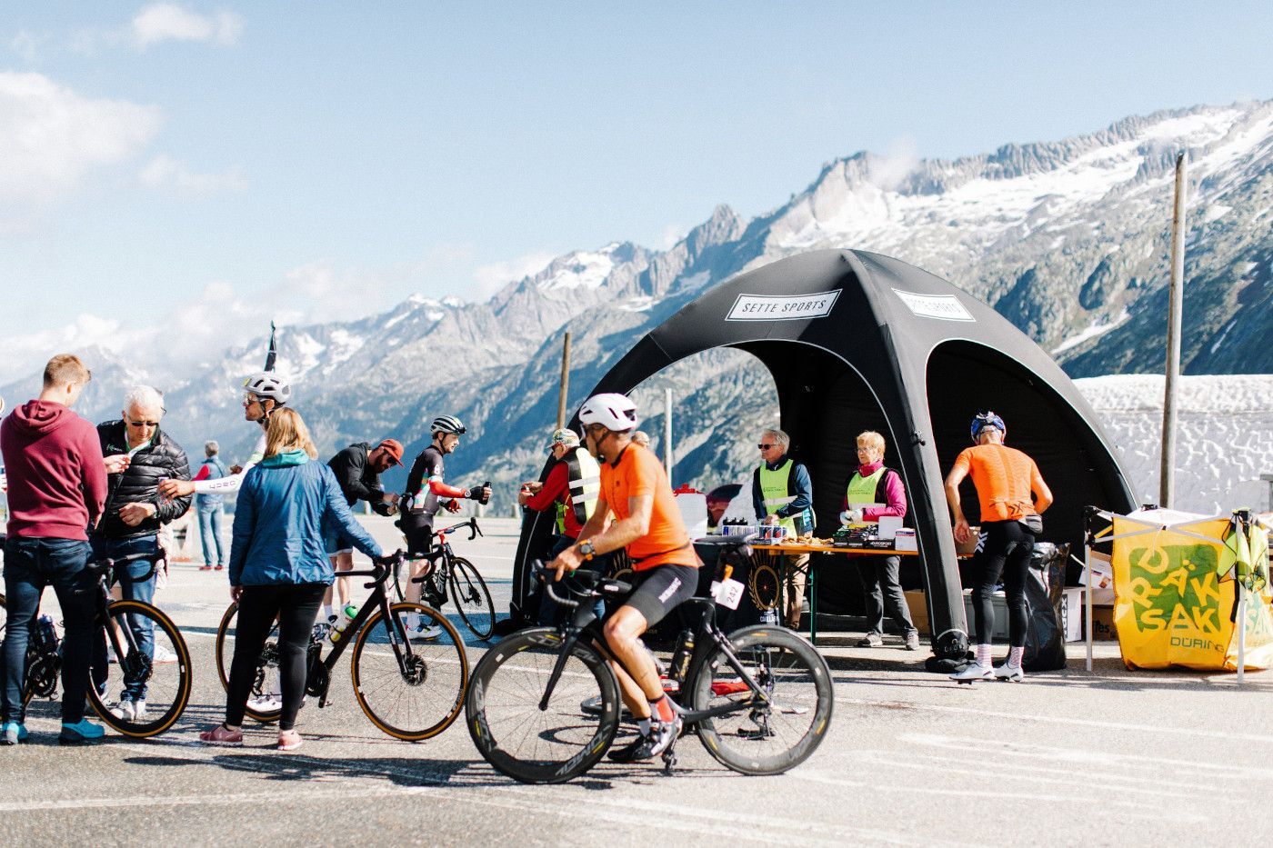 Cyclists gathered at a mountain overlook with snowy peaks and a black event tent in the background