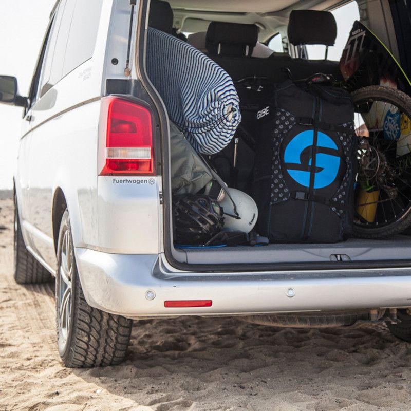 White van parked on sand with the rear hatch open, packed with luggage and gear
