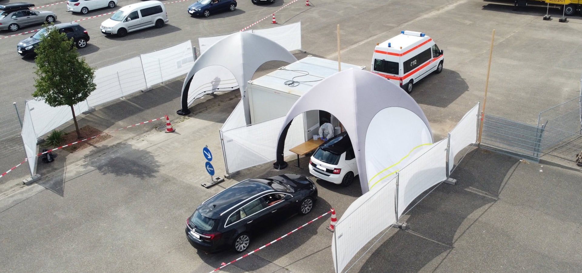 Cars entering a white tented drive-through checkpoint in a parking lot, with an ambulance nearby.