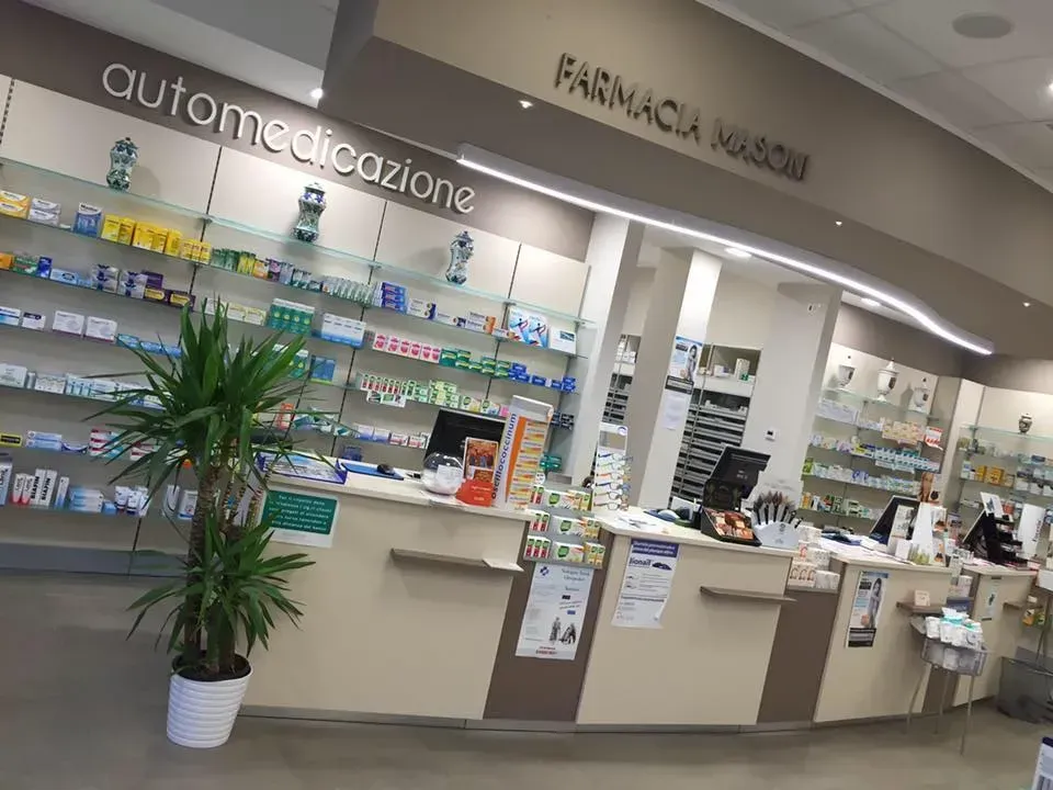 Interno della farmacia Masoni: scaffali pieni di medicinali, bancone con personale, pianta in vaso davanti.