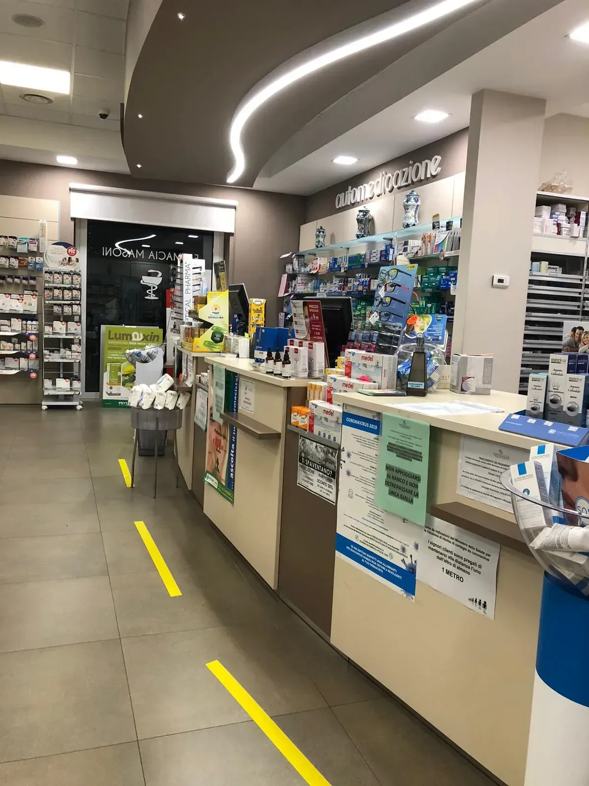 Interno della farmacia Masoni con bancone, prodotti e segnaletica gialla sul pavimento.