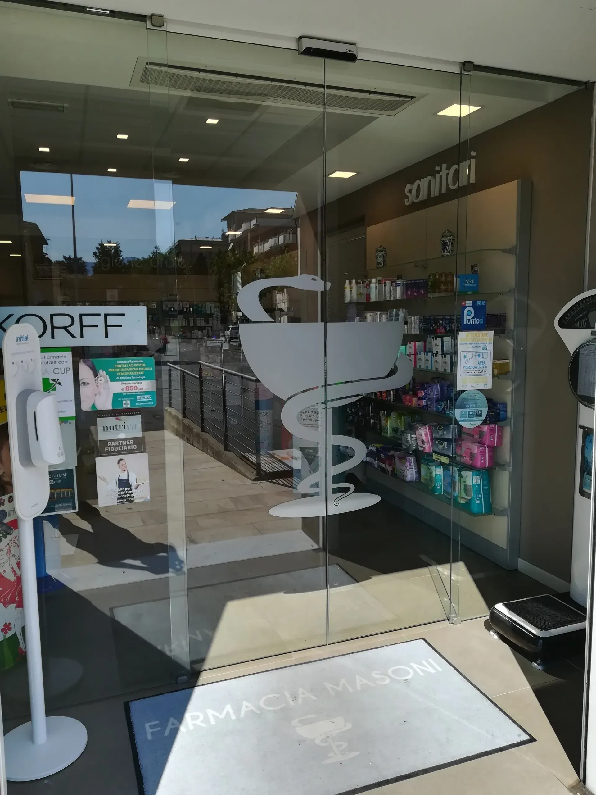 Ingresso della farmacia Masoni con porta in vetro con il simbolo della farmacia, insegna Sanicar e dispenser di disinfettante per le mani.