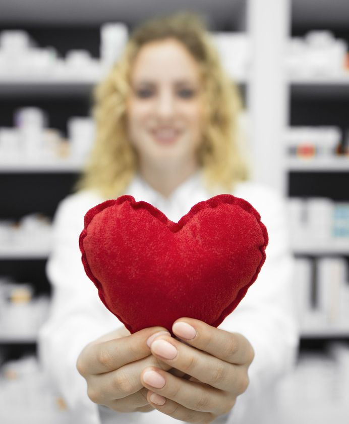 Una donna in camice bianco tiene in mano un cuore rosso; sullo sfondo, scaffali di medicinali.