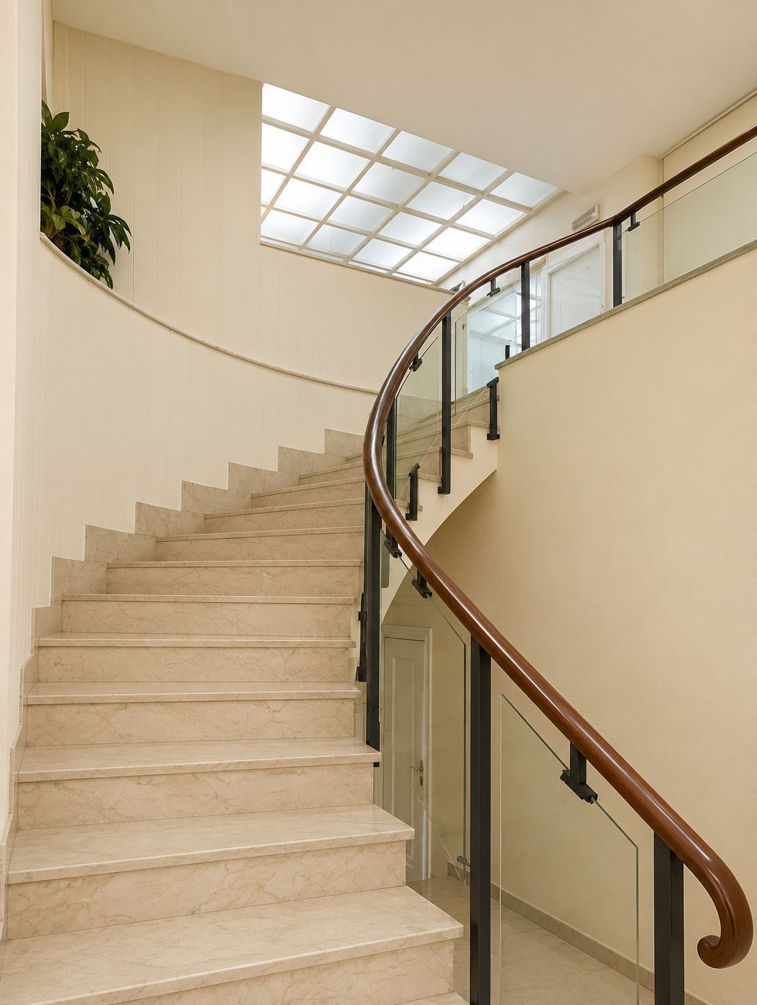 Escalera interior curva con paredes beige, barandilla de cristal y tragaluz en la parte superior.