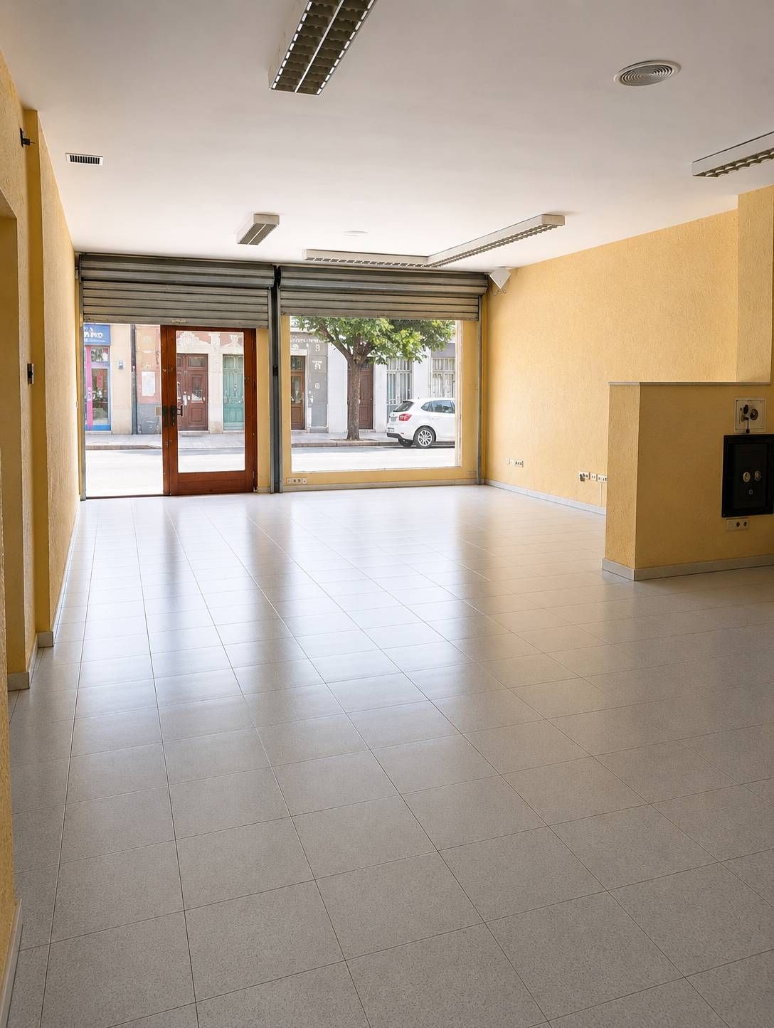 Interior de un local comercial vacío con paredes de color beige, suelo de baldosas y grandes ventanales que dan a la calle.