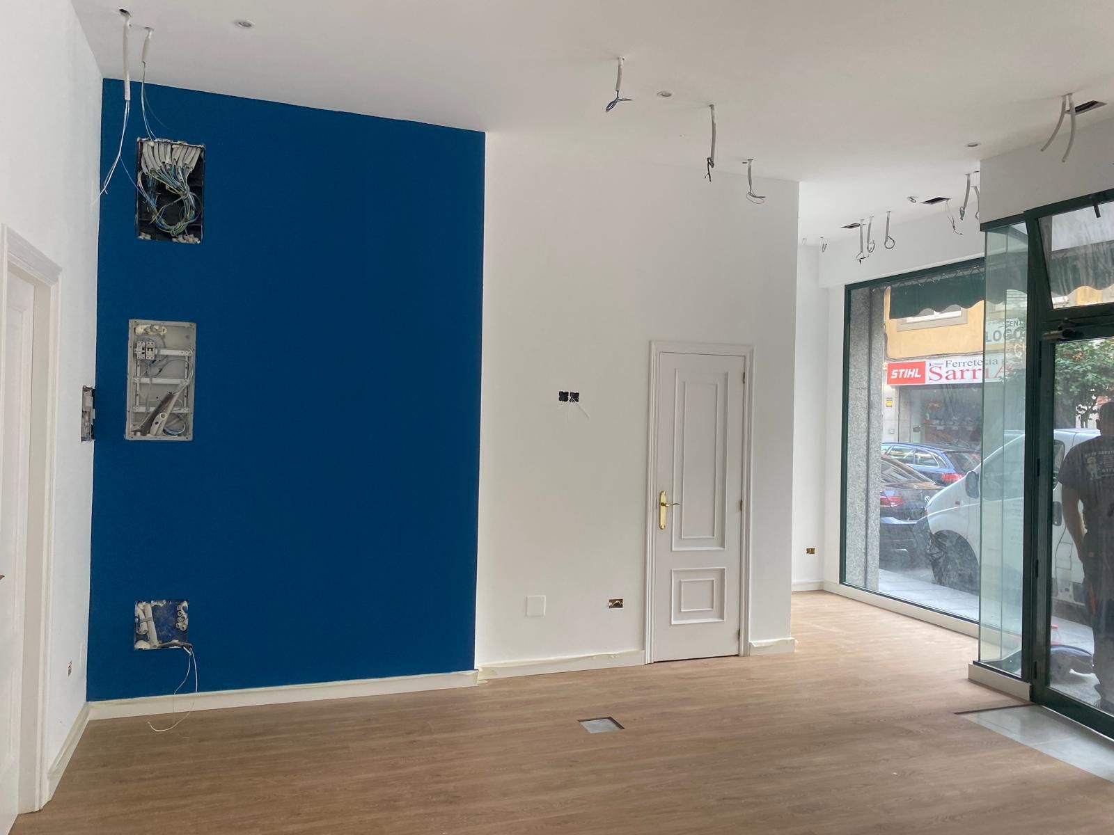 Interior de local comercial vacío con pared decorativa azul, suelo de madera, puerta blanca y grandes ventanales.
