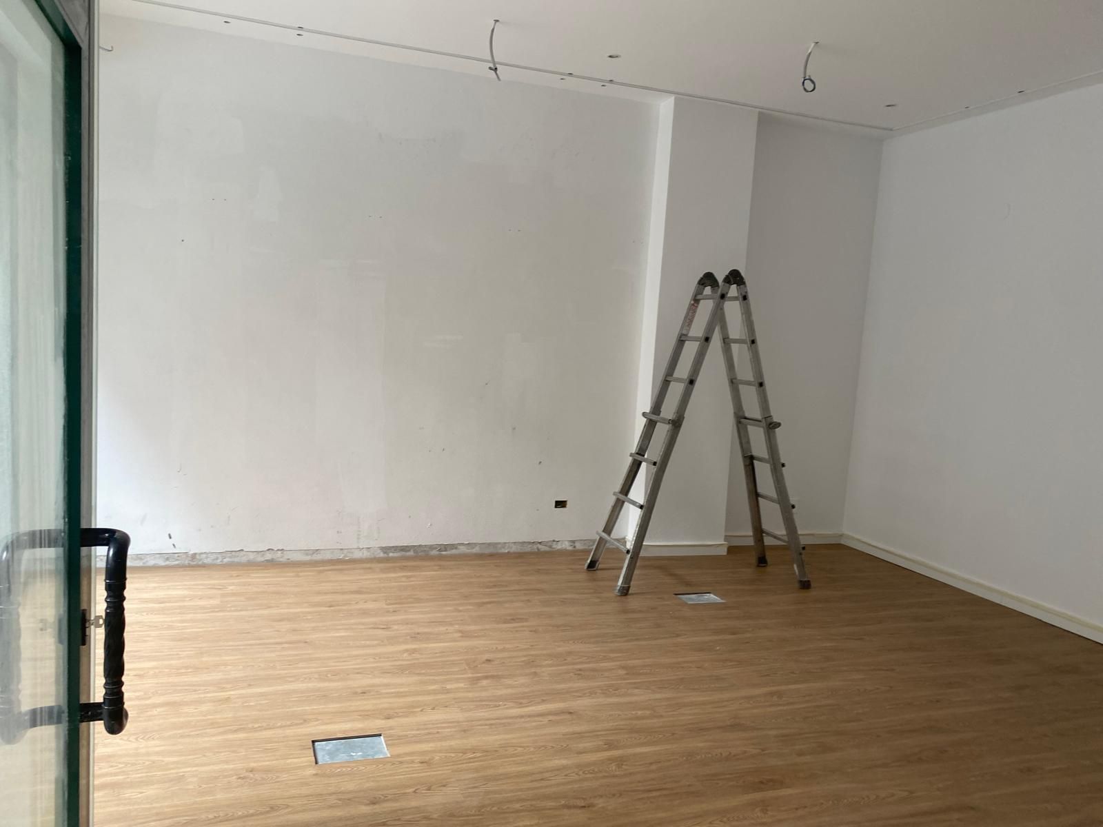 Habitación vacía con paredes blancas, suelo de madera y una escalera abierta cerca de la pared del fondo.