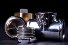 Accesorios de tubería metálicos y un conector roscado dispuestos sobre una superficie negra sobre un fondo oscuro.