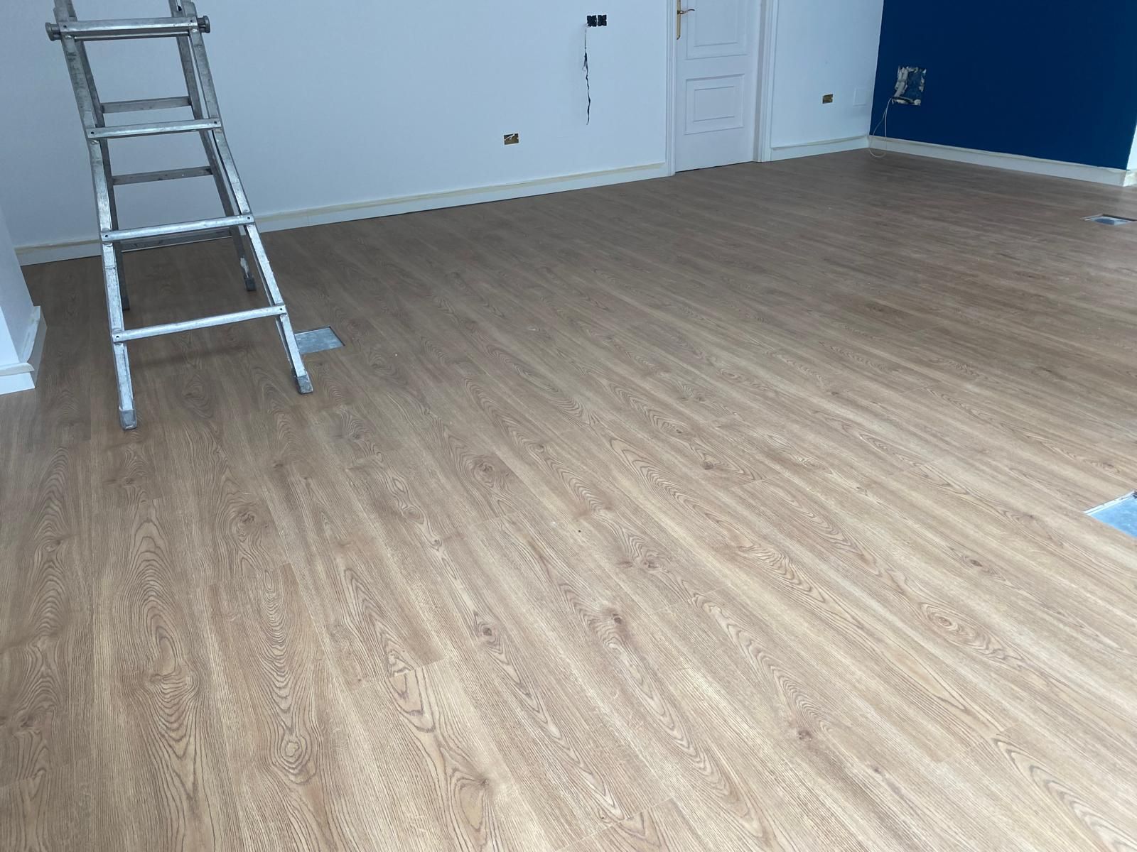 Habitación vacía con suelo de madera clara, una escalera de mano, paredes blancas y una pared decorativa de color azul.