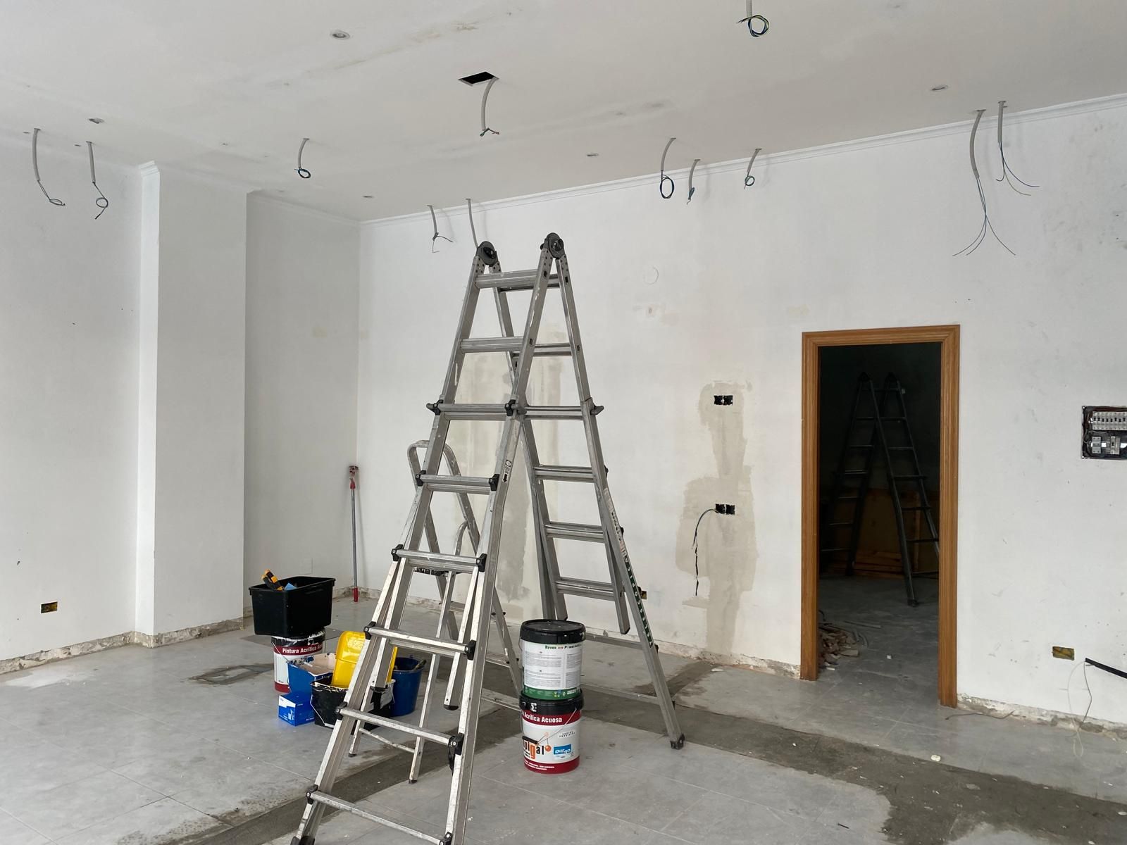 Cuarto de construcción con escaleras, cubos de pintura y paredes blancas sin terminar, con cables expuestos en el techo.