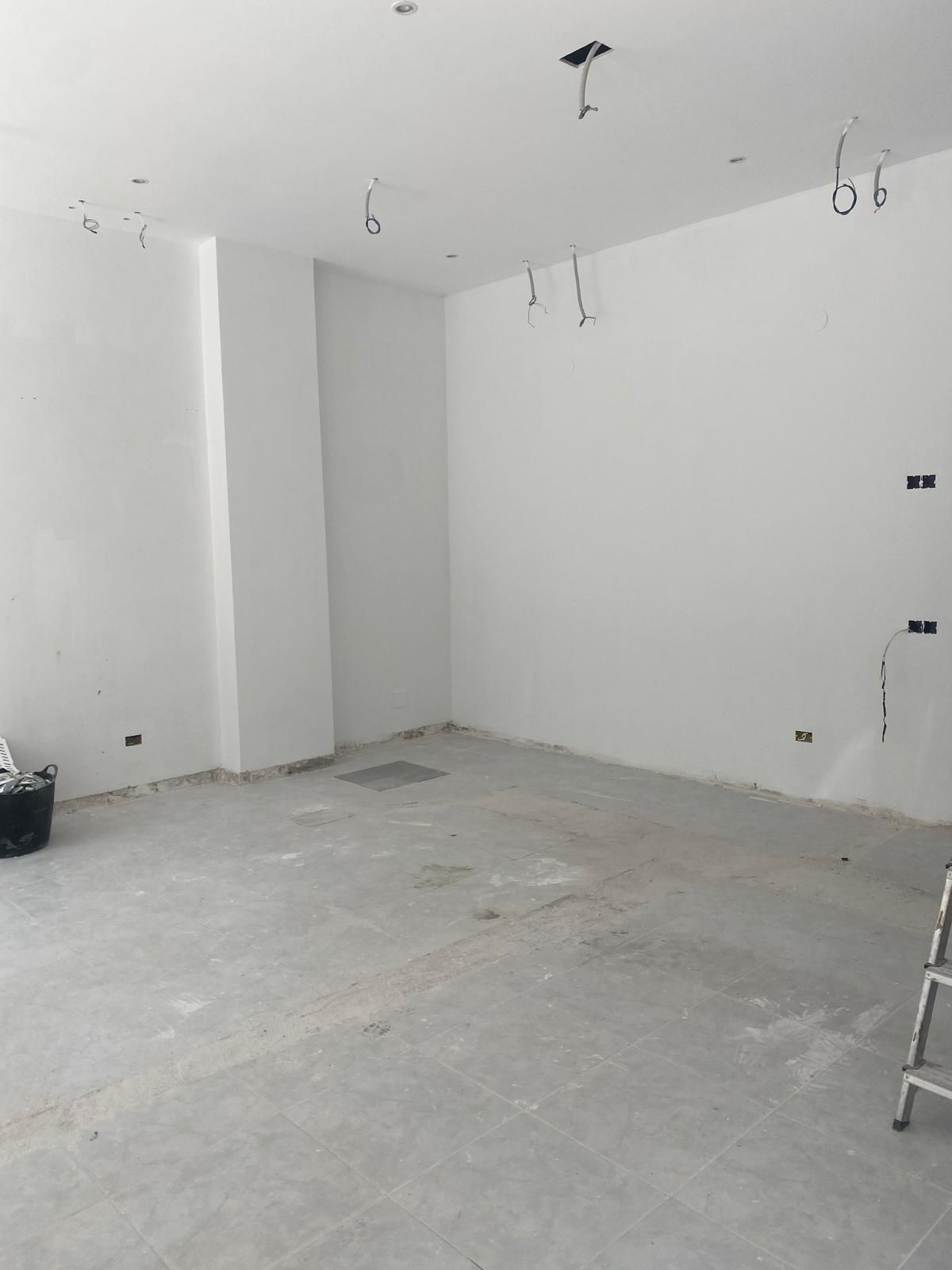 Habitación vacía sin terminar con paredes blancas, suelo de hormigón visto y cableado expuesto en el techo.