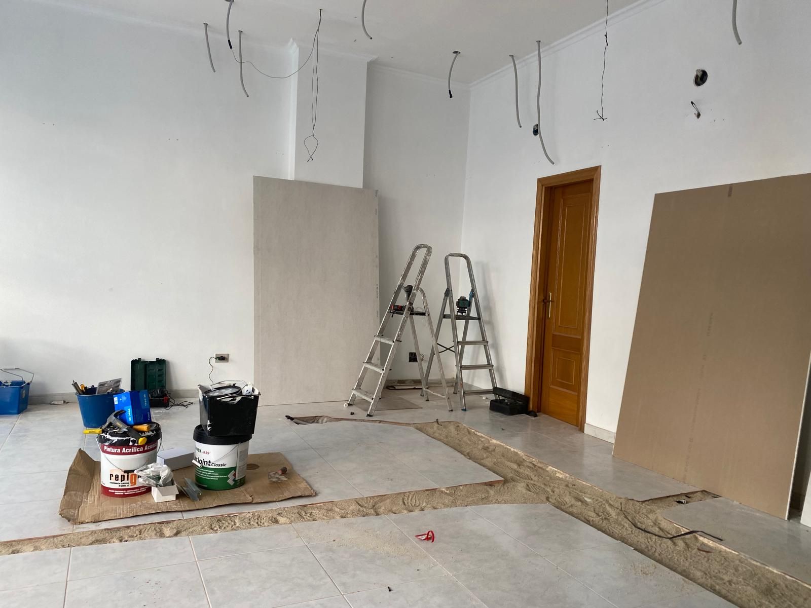 Habitación parcialmente terminada con escaleras, cubos de pintura y cableado expuesto en paredes blancas.