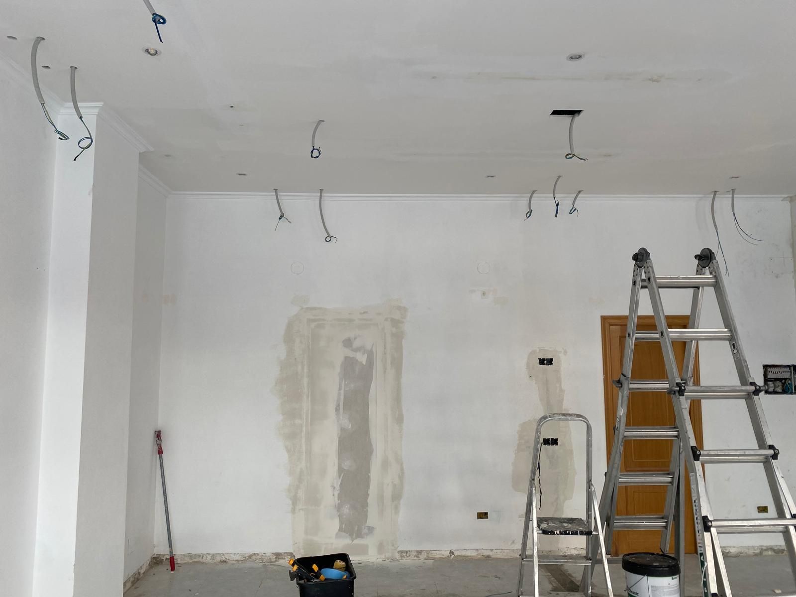 Habitación sin terminar con cableado del techo a la vista, escalera y materiales de pintura sobre paredes blancas.