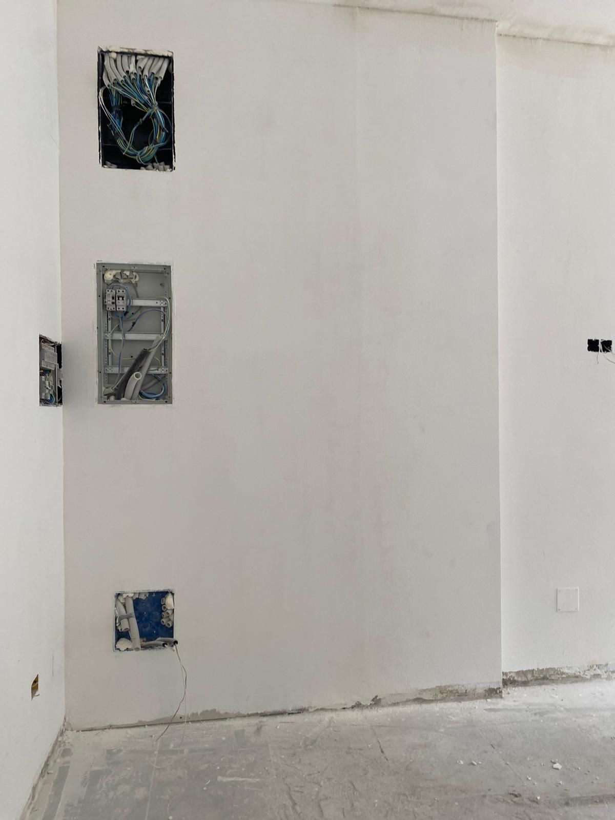 Pared interior blanca vacía con cajas eléctricas a la vista y suelo sin acabar.