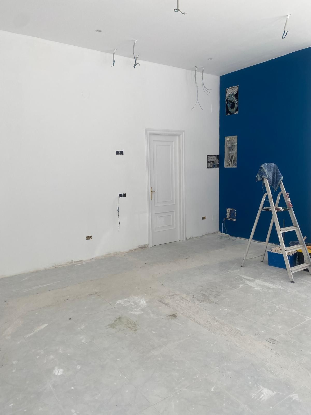 Habitación vacía en obras, con paredes blancas y azules, escalera y suelo a la vista.