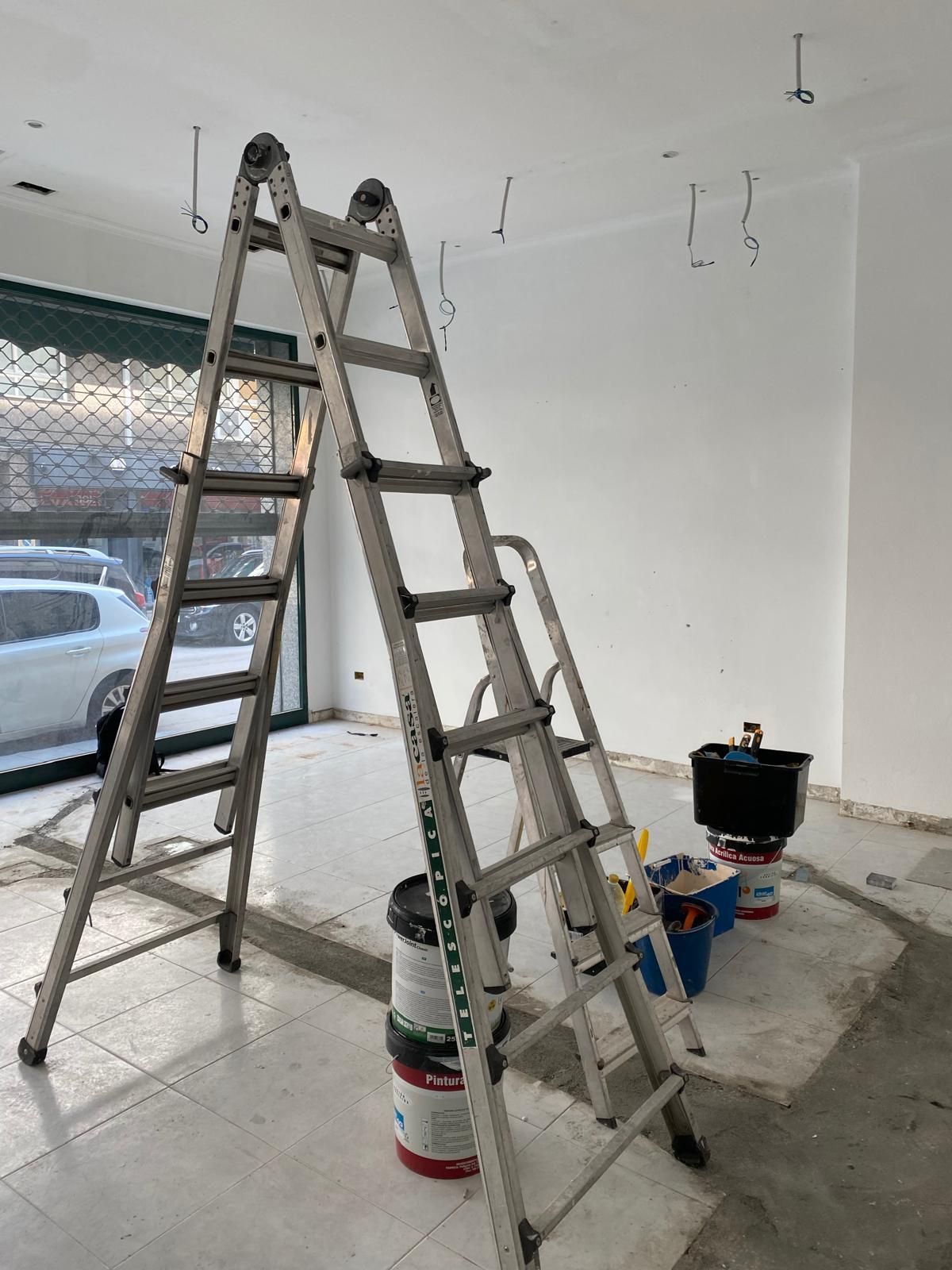 Dos escaleras en una habitación casi vacía con latas de pintura y herramientas en el suelo.