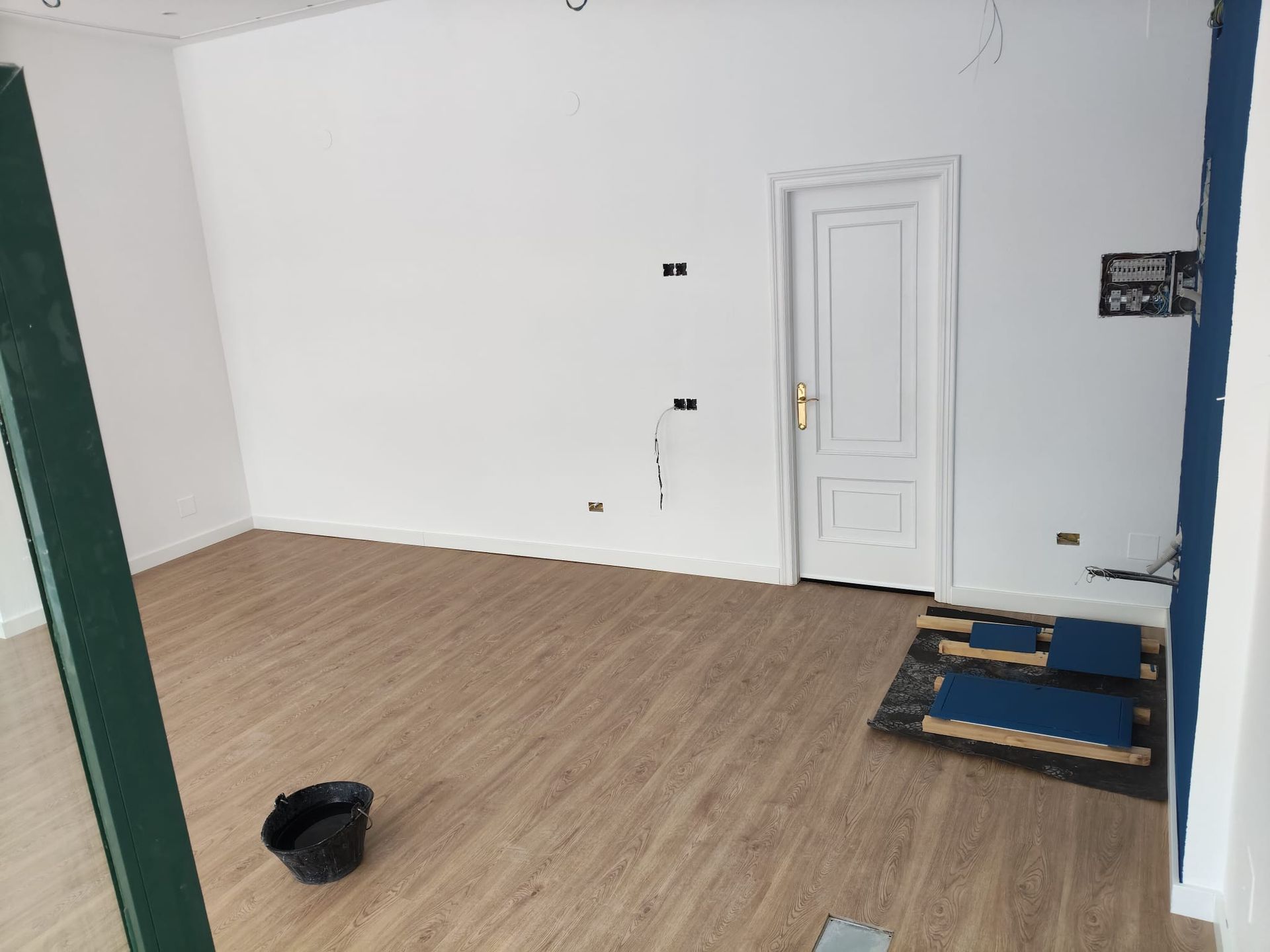 Habitación vacía con suelo de madera, paredes blancas y puerta cerrada; algunos objetos yacen en el suelo cerca de la pared derecha.