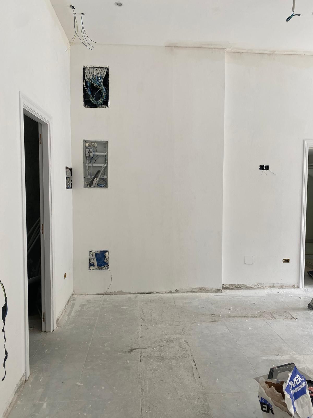 Habitación blanca vacía en proceso de renovación con suelo desnudo, aberturas en las paredes y materiales de construcción esparcidos por todas partes.
