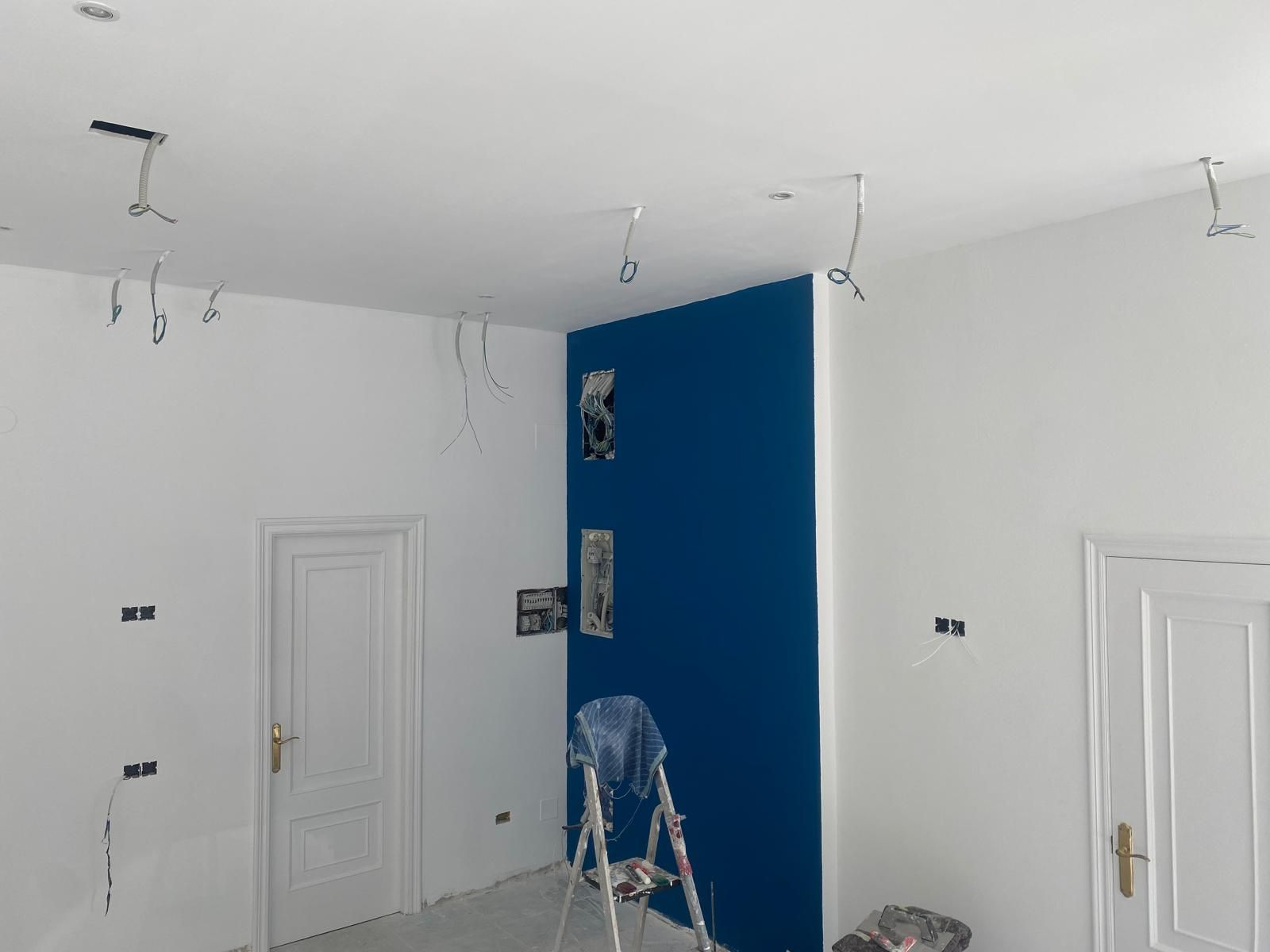 Habitación blanca en proceso de renovación con pared decorativa azul, escalera y cableado en el techo.