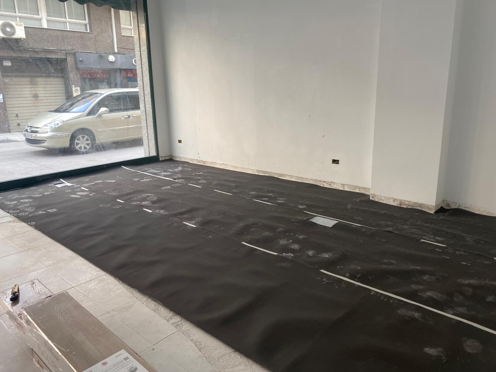 Local comercial vacío con suelo negro, paredes blancas y una ventana con vistas a coches aparcados.