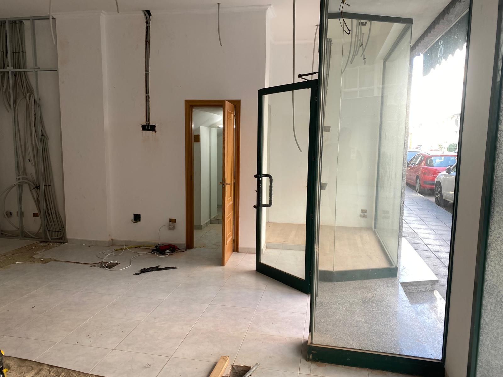 Habitación vacía sin terminar, con puerta de cristal abierta, paredes blancas y suelo de hormigón, cerca de una acera.