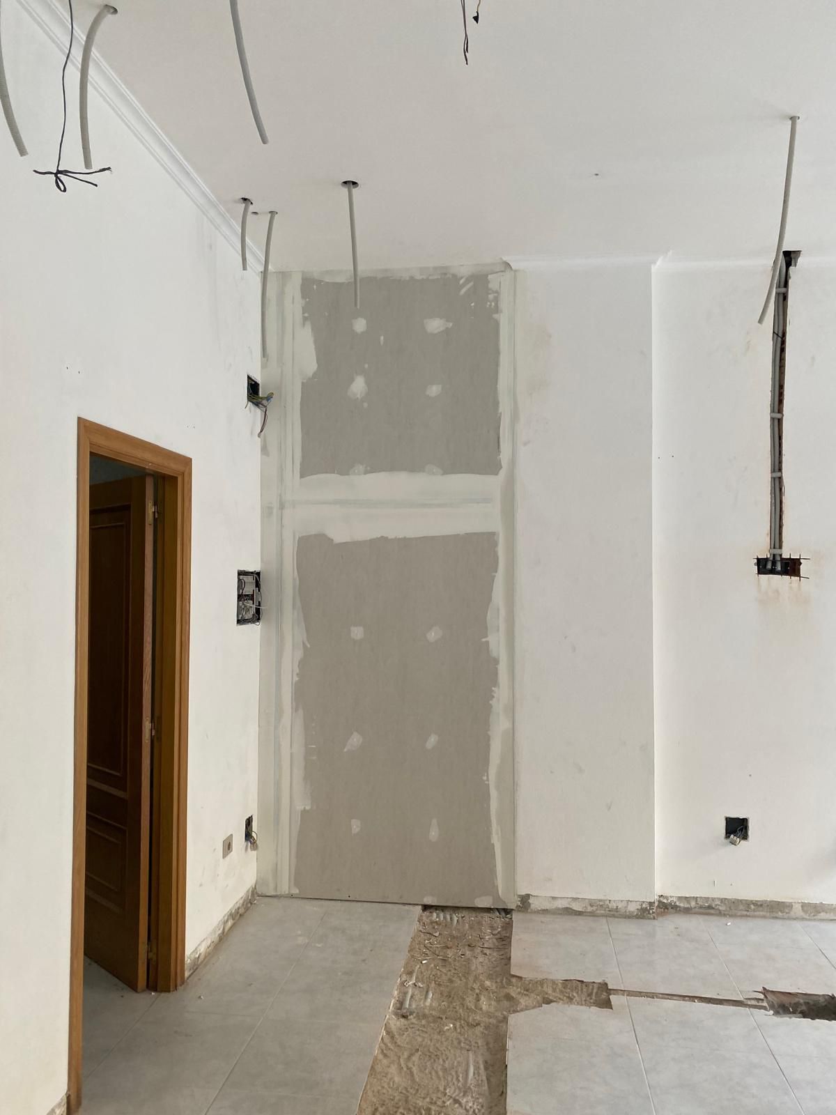 Habitación interior blanca y vacía con parches de paneles de yeso sin terminar y cableado expuesto cerca del techo.