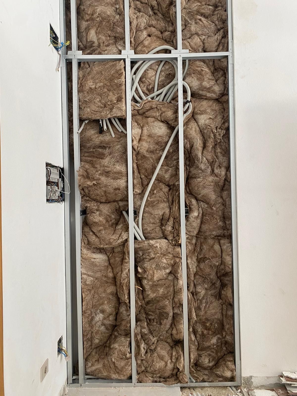 Cavidad de pared abierta con aislamiento marrón expuesto detrás de un marco de metal blanco.