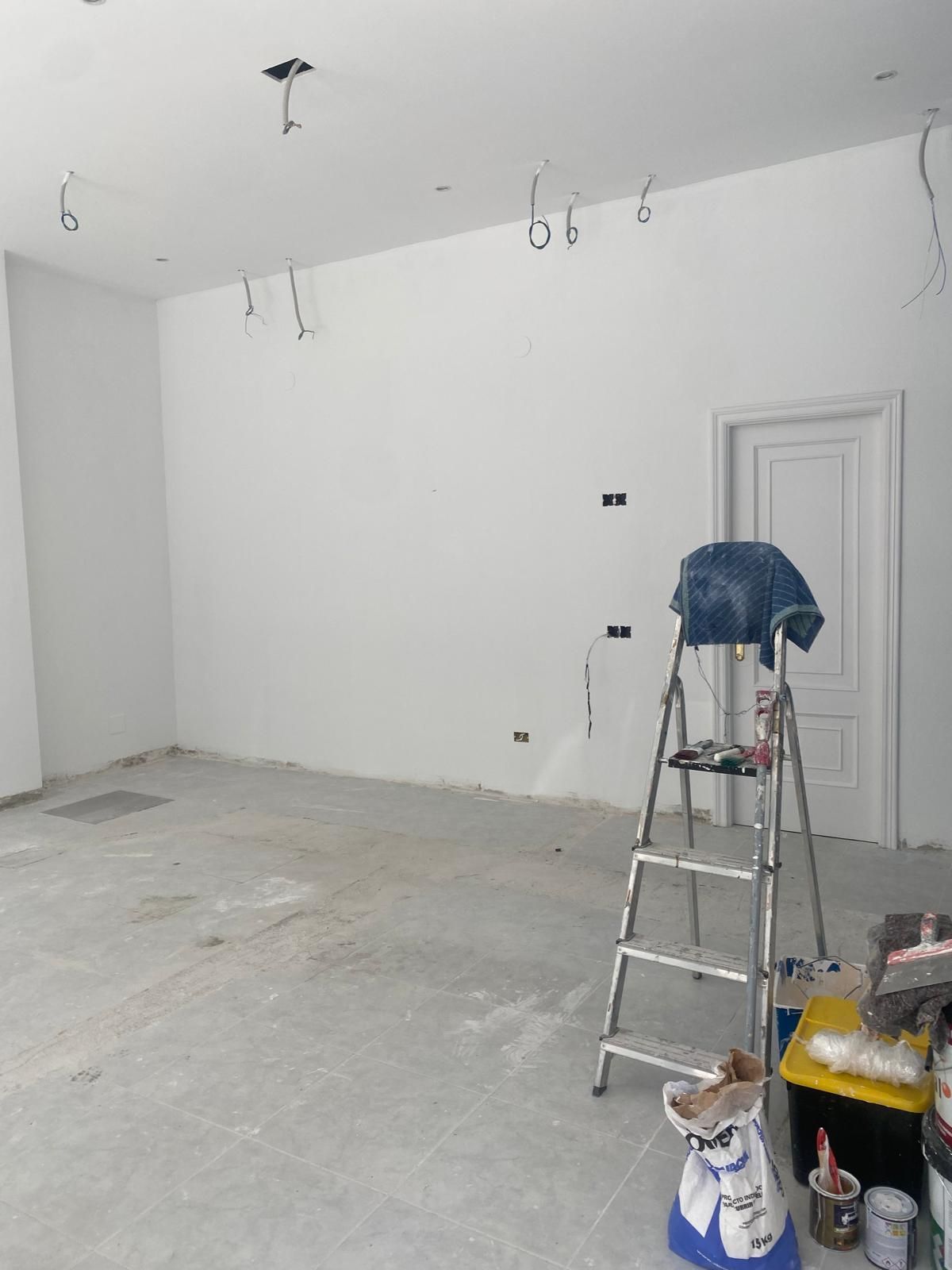 Habitación vacía en obras, con paredes blancas, suelo desnudo y una escalera de mano con herramientas.